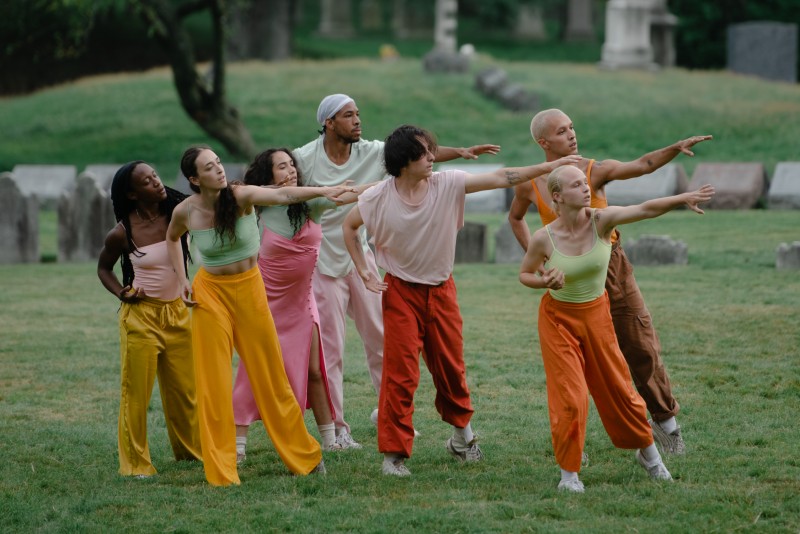 Seven dancers stand in a clump with one arm extended, the other bent at their sides.